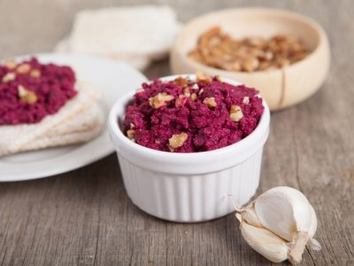 Beetroot pesto in a white bowl on a wooden table with garlic beetroot and rice cakes on the back, selective focus