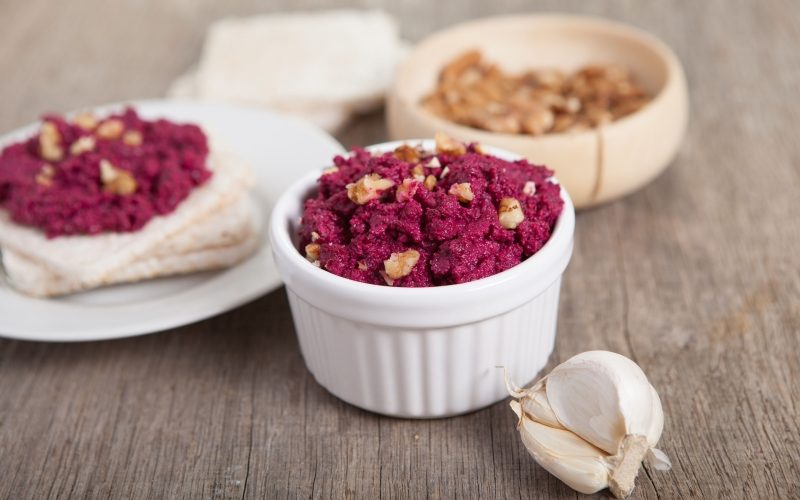 Beetroot pesto in a white bowl on a wooden table with garlic beetroot and rice cakes on the back, selective focus