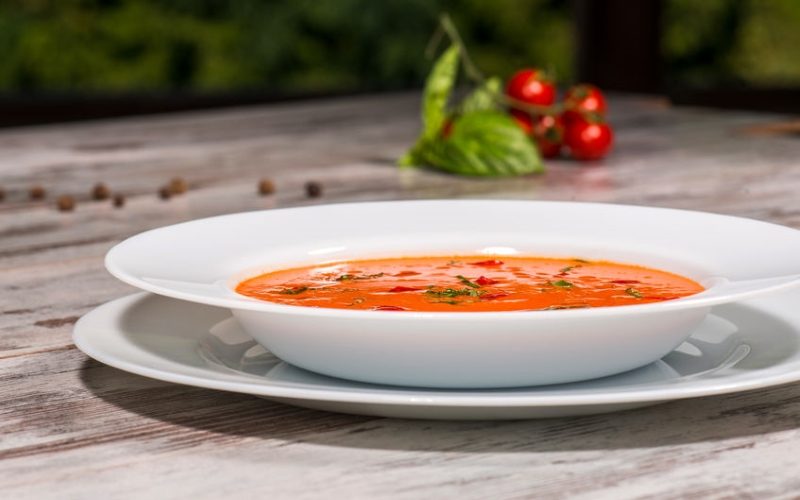 Close-up low-angle picture of cold tasty tomato soup gazpacho with species on the wooden table in vegan cafe with tomatoes standing near a plate, with copy place and selective focus on a plate